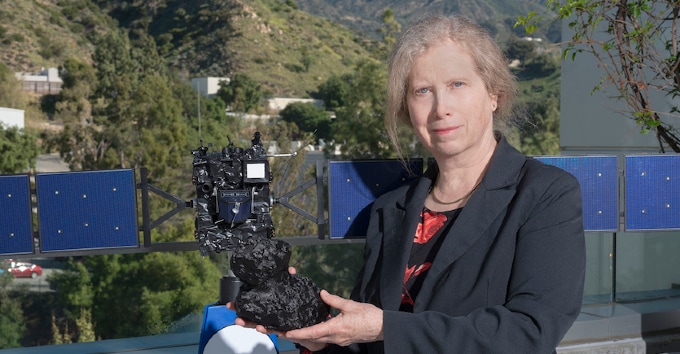 An elderly white woman in a black blazer standing in front of a model of a spaceship and holding a model of an outer solar system object.